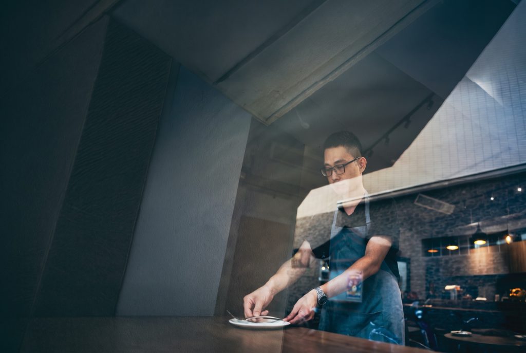 A restaurant worker in glasses and an apron sets down a plate, seen through the window